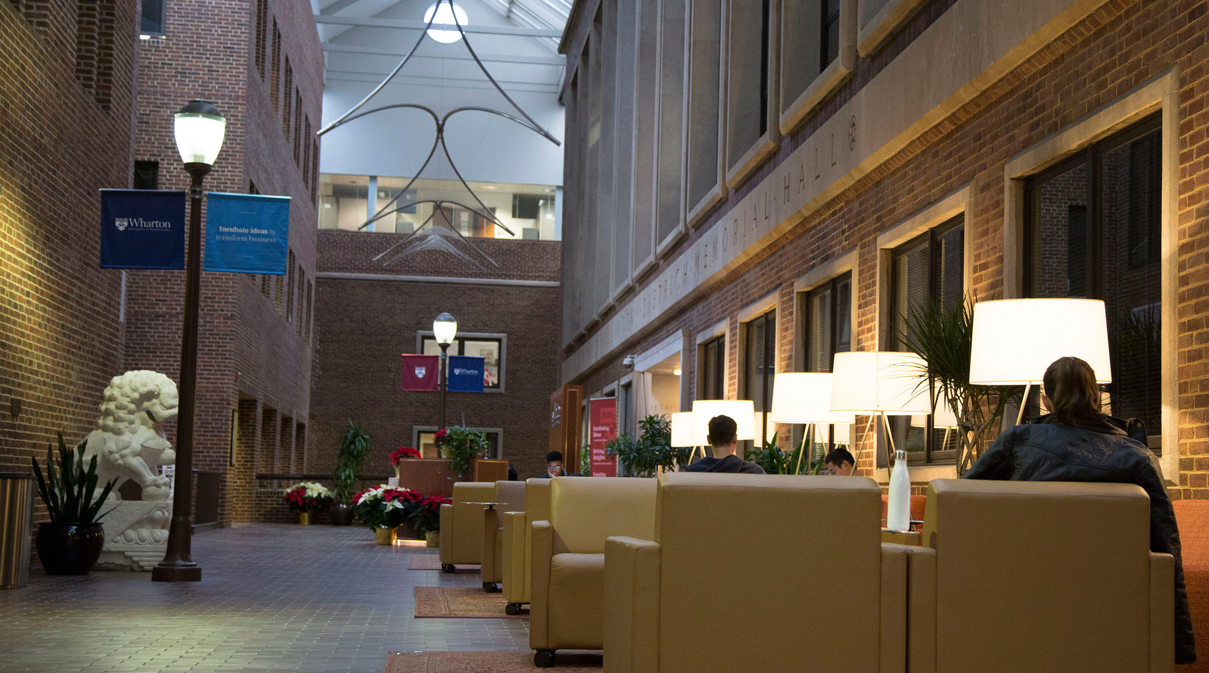 A modern indoor seating area with beige armchairs, floor lamps, and people studying. Large windows, brick walls, and banners with "Wharton" are visible.