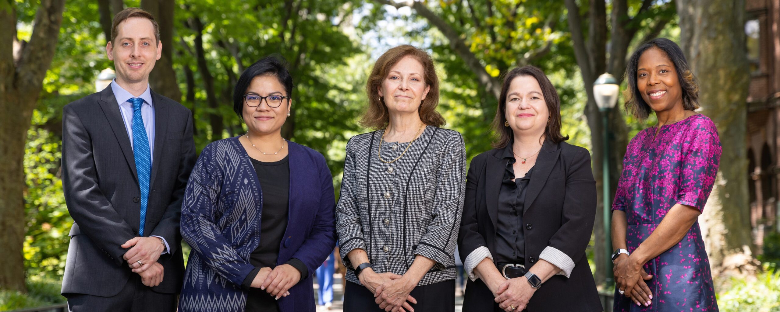 A group photo of five people standing outdoors in a leafy setting. They are dressed in professional attire, with trees and a lamp post in the background.