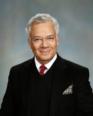 Headshot of a person with short, gray hair, wearing glasses, a black suit with a red tie, and a patterned pocket square, against a neutral background.