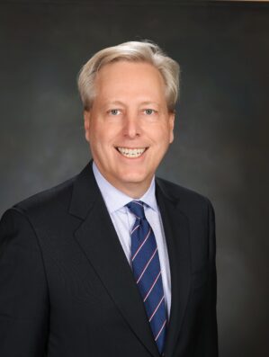 Headshot of a person wearing a suit and tie, smiling against a dark, neutral background.