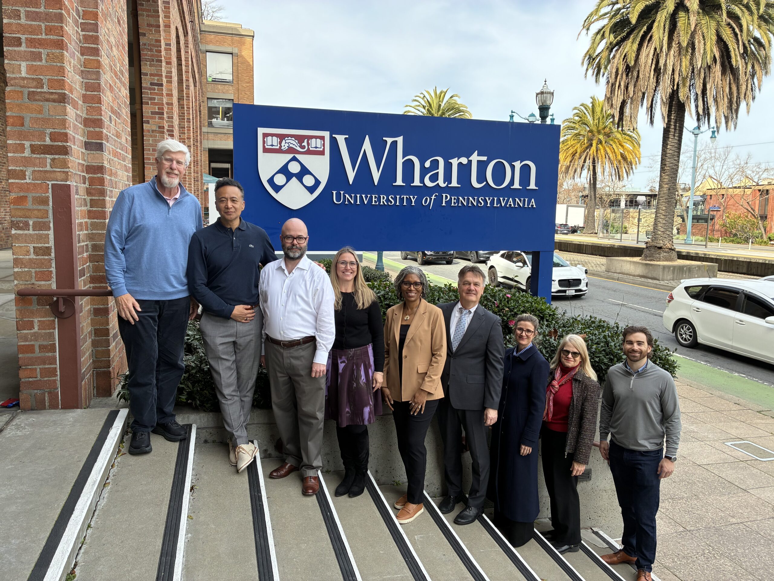 A group of people standing in front of a Wharton School of the University of Pennsylvania sign, outdoors with palm trees and a building in the background.